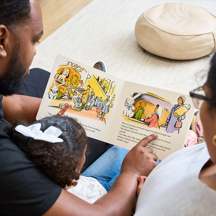 A family of three reads a children's book together, with the child sitting in the lap of one adult while the other two concentrate on the book's illustrations and text.