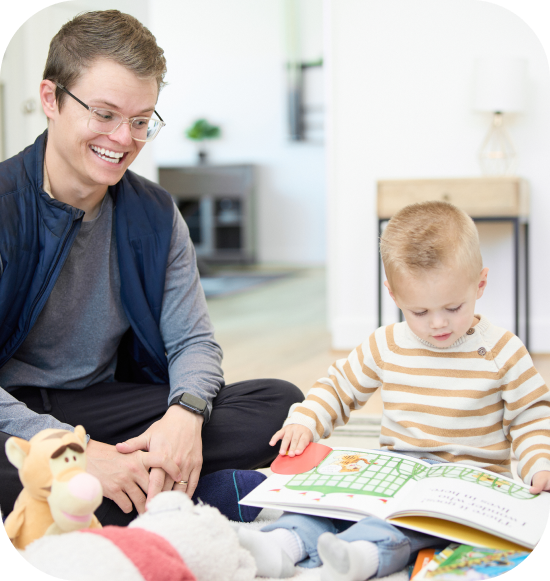 An adult and a child sit on the floor among toys while the child looks at an open book.