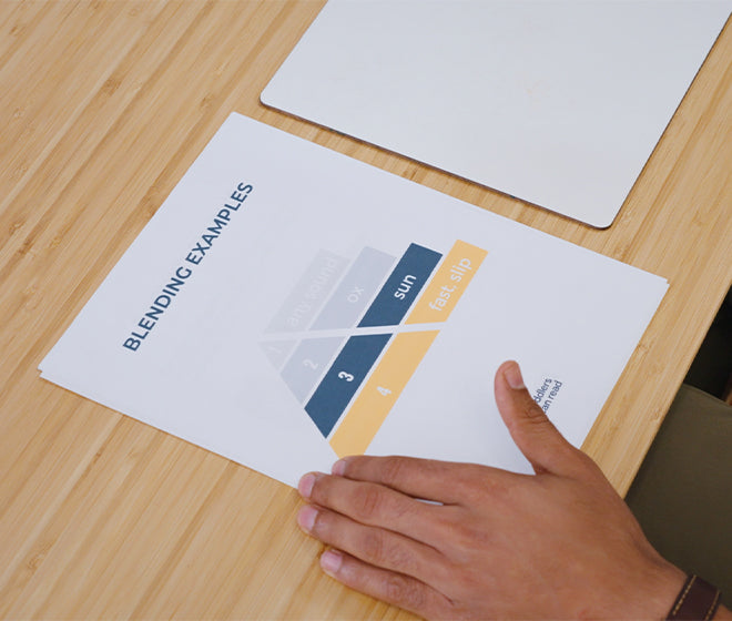 Person's hand holding a paper titled Blending Examples on a wooden desk, showcasing a color blending chart with labels 1-4 and colors in blue, gray, yellow, and peach.