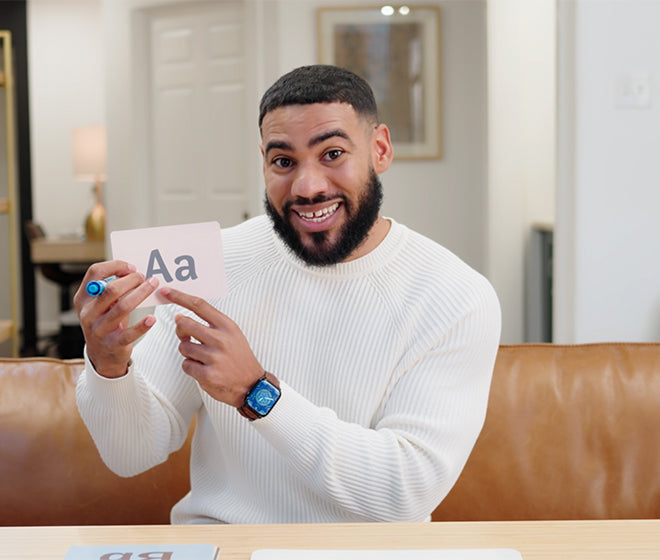 A man in a white sweater holding a flashcard with the letters Aa while sitting on a brown sofa.