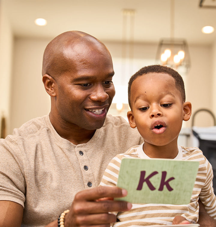 A man and a young boy look at a card with the letter Kk printed on it. The boy's mouth is open, as if he is reading or pronouncing the letter. They are seated in a brightly lit room.