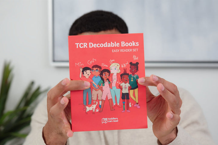 Person holding up a red book titled TCR Decodable Books: Easy Reader Set featuring illustrations of five diverse children and a dog on the cover.