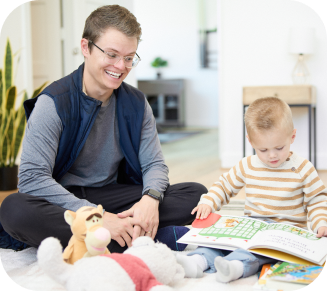 An adult and a child sit on a rug, reading a book. Various toys are placed near them. The adult is smiling and sitting cross-legged, while the child is focused on the book.