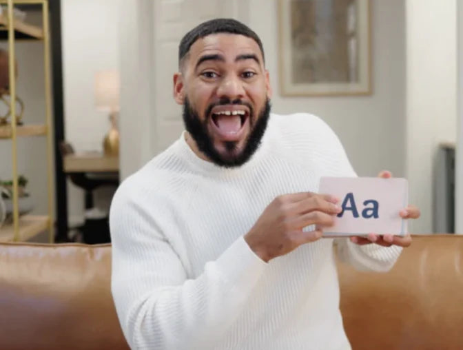 A man with a beard and a white sweater holds up a sign with Aa on it while sitting on a sofa, smiling enthusiastically.